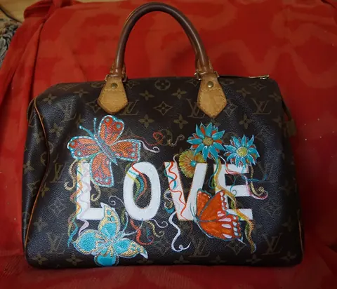 Sold original by Agata May'kowska — A brown Louis Vuitton handbag with floral and butterfly embroidery and the word "LOVE" on the front, resting on a red fabric surface.