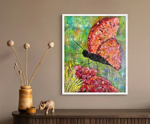 Sold original by Agata May'kowska — Colorful abstract painting of a butterfly with orange and pink wings, framed white, on a beige wall.