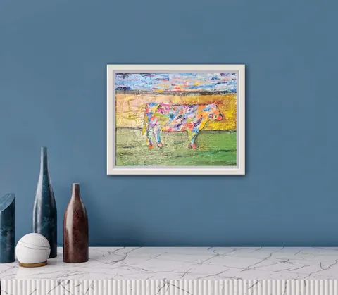 Sold original by Agata May'kowska — Colorful framed painting of a cow against a landscape background, hung on a blue wall above a white marble table with decorative vases.
